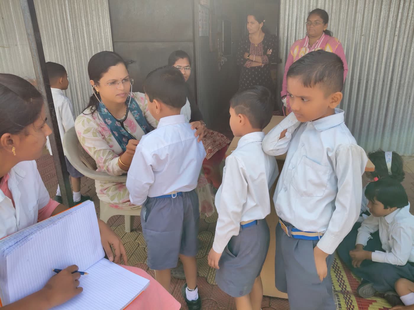 A child being checked with a stethoscope while a teacher takes notes and other children wait in line