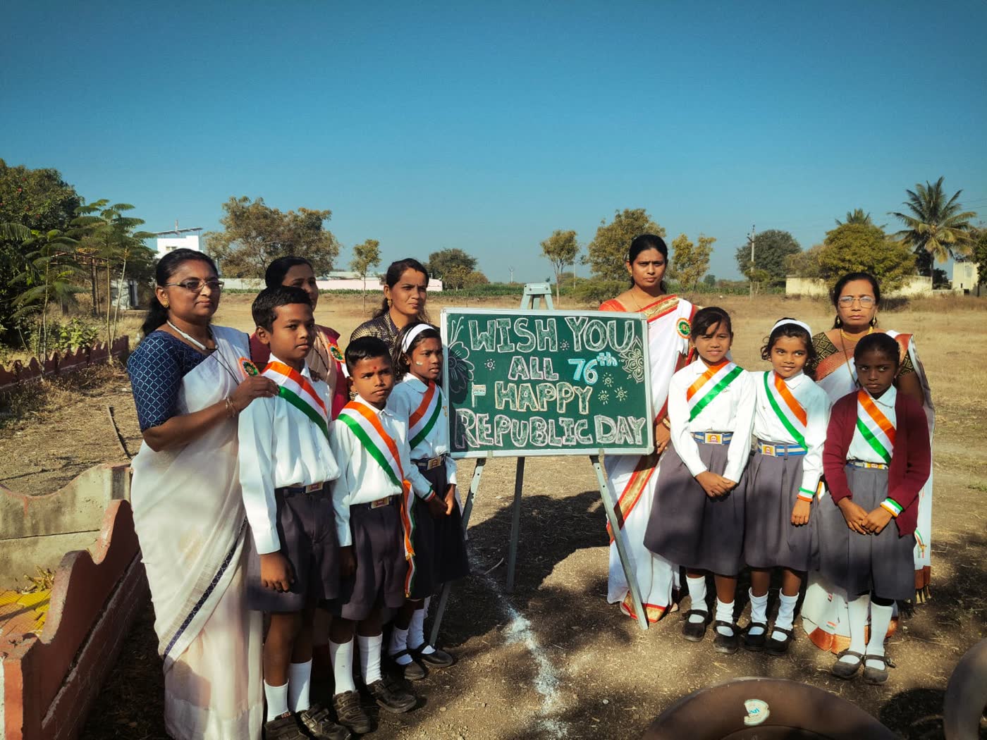 Students with tricolour sashes and their teachers beside a green board reading Wish you all 76th Happy Republic Day