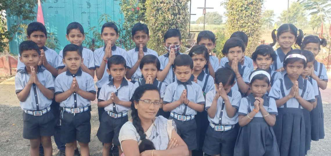 Children in school uniform with their hands folded in greeting, their teacher kneeling in front of them
