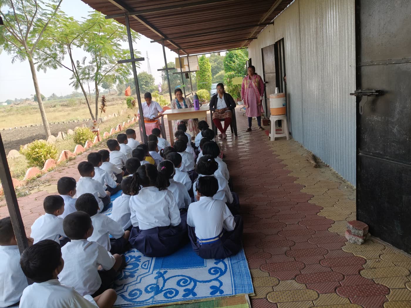 Students seated on mats in the school verandah, listening to their teachers